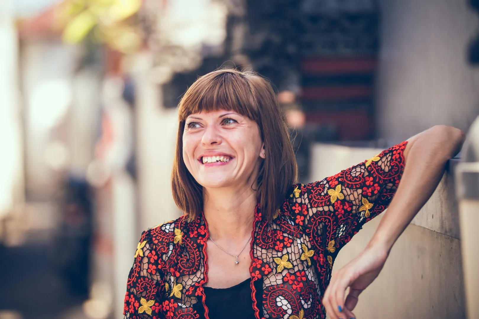 Smiling woman with a fringe wearing a red embroidered lace top, leaning on a wall outdoors