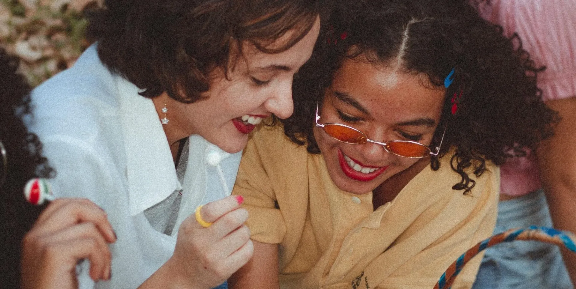 Two young women laughing together, one holding a lollipop and wearing tinted glasses