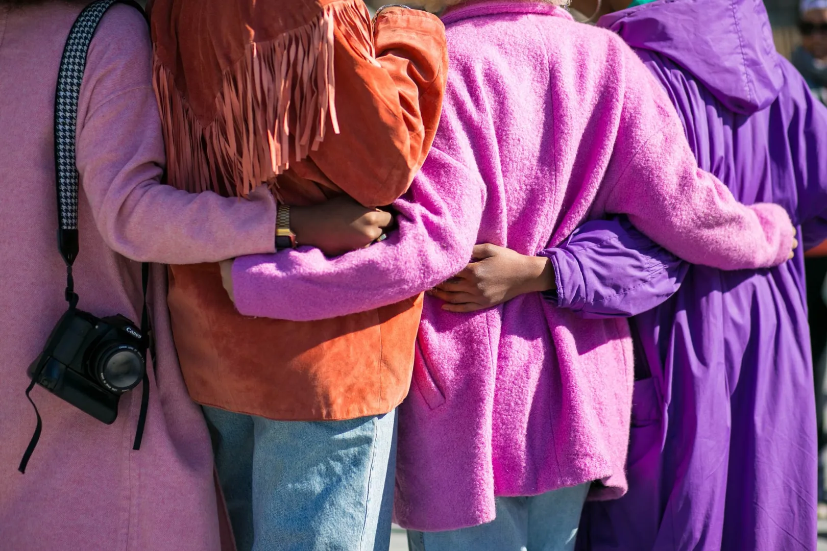 Four friends in pink and purple jackets standing arm in arm, seen from behind
