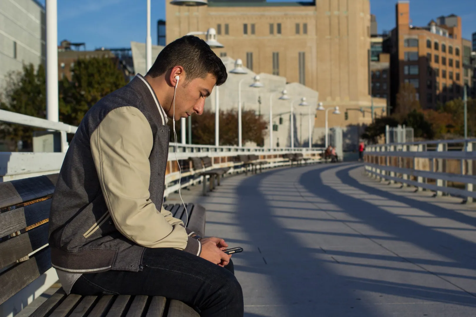 Young man wearing earphones looking at his phone on a bench on a New York riverside walkway
