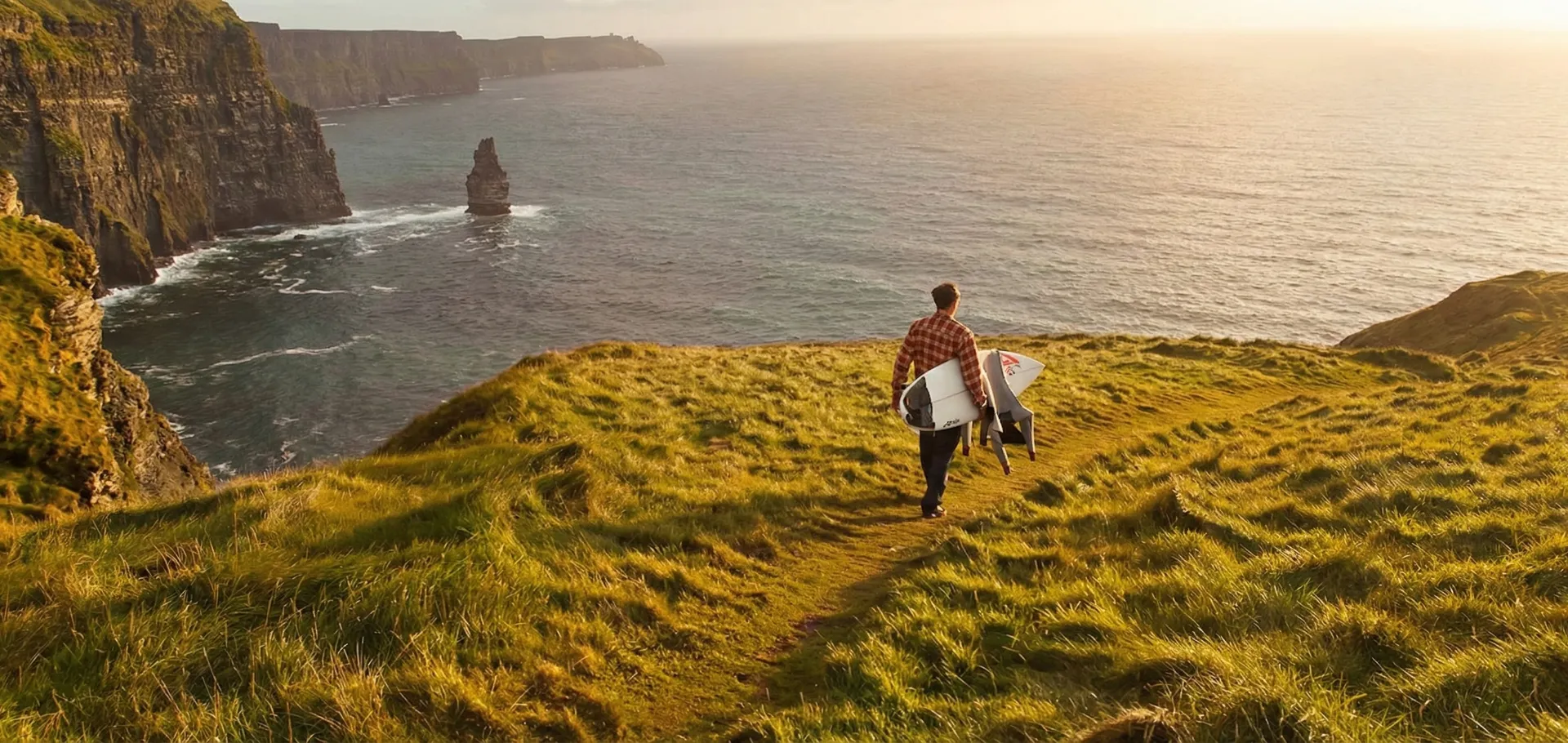 Surfer carrying a board along a grassy clifftop path above the sea at golden hour