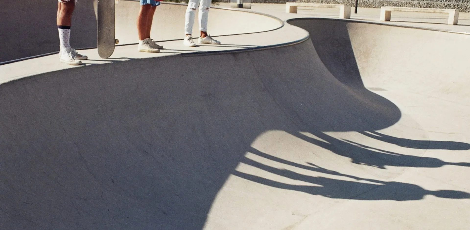 Teen boys standing with a skateboard at the edge of a sunlit concrete skate bowl